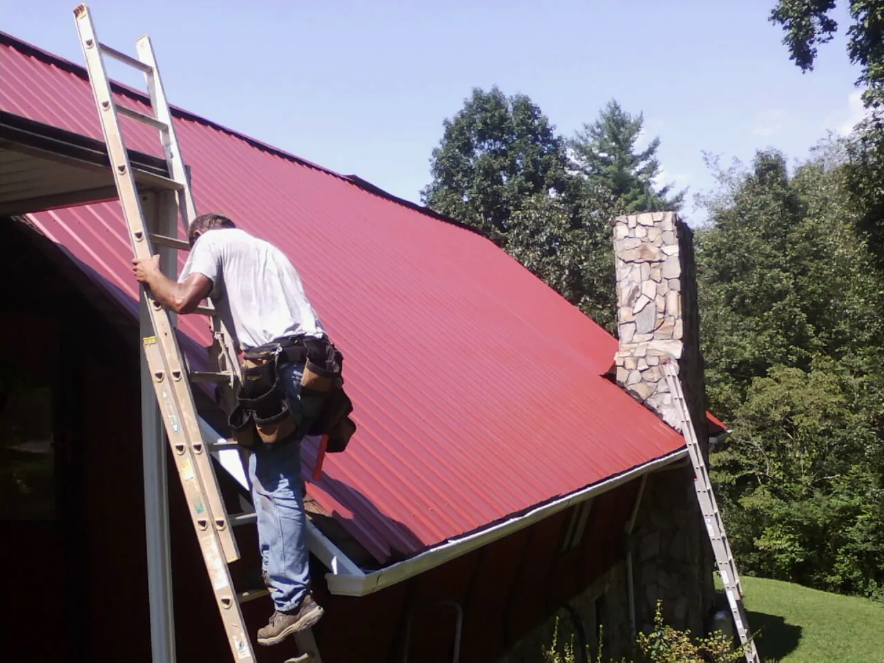 Roofer on a ladder beside a red metal roof and stone chimney — Soffit & Fascia Repair by Roofing Brothers in Nicktown